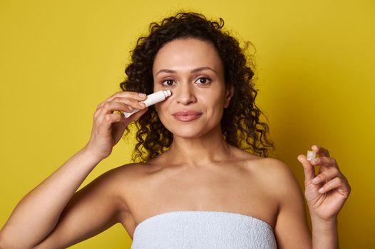 Close-up portrait of an attractive natural beauty woman wrapped in a white bath towel holding a tube of cosmetic product and applying it under the eyes, isolated over yellow background with copy space
