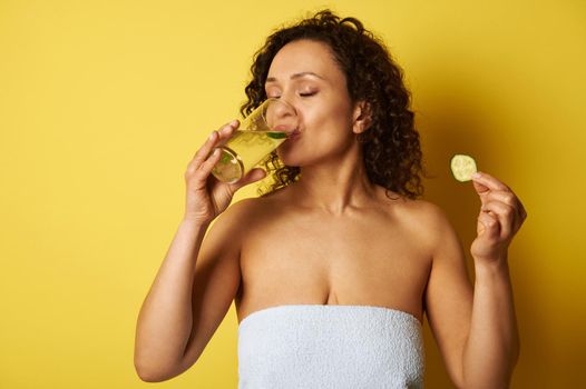 Young half-naked woman, wrapped in a towel, holding a slice of cucumber and drinking water from a glass, standing against a yellow background with space for text