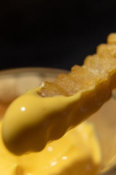 Close-up of wavy French fry being dipped into glass of melted cheddar cheese with black background. Fried potato snack covered with tasty yellow dip. Delicious fast food preparation