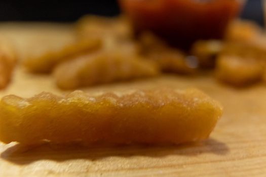 Extreme close-up of wavy French fry on wooden surface with blurry cup of ketchup as background. Tasty fried snacks and glass of red tomato dressing. Delicious fast food