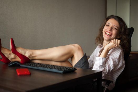 Portriat of brunette with legs on office table with laptop, hot woman in red shoes on high heels sitting at the desk. Concept of seductive secretary, female fashion, relax at work.