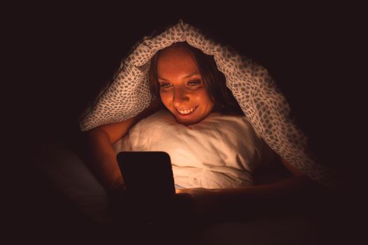 Happy smiling woman lying in bed under the blanket and typing on smartphone at night before sleeping. Social media addiction and communication. High quality photo