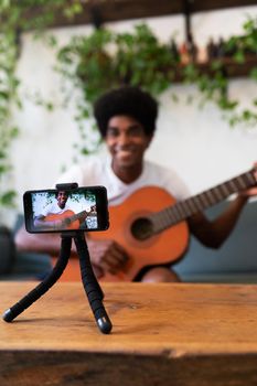Smiling young african american man recording a guitar online lesson with mobile phone. Selective focus on phone. Vertical image. E-learning concept.
