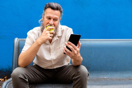 Mature caucasian man using mobile phone and eating an apple in a public park. Copy space. Lifestyle concept.