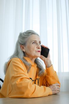 Grandmother with grey hair and face with wrinkles is using smartphone, talking with someone and sitting at the table at home on window background, mother's day, happy retirement, vertical