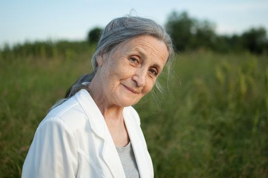 Portrait of senior woman with grey hair and face with wrinkles wearing white jacket and relaxing at park during sunny day, happy retirement, mothers day concepts