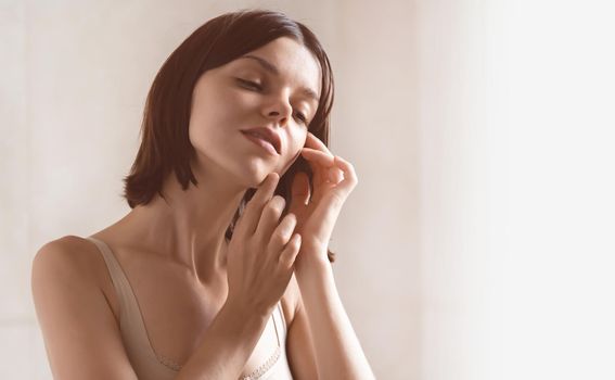 A young girl touches her delicate, clean, moisturized face skin with her hands, a woman takes care of the health and beauty of her body in the bathroom.