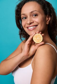 Side portrait of attractive delighted woman holding a half of yellow fresh juicy lemon near her face and smiling with beautiful healthy toothy smile posing against bright blue background Copy ad space