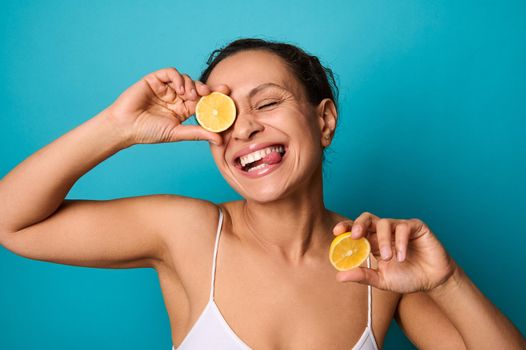 Beauty portrait of attractive cheerful smiling young woman with curly dark hair wearing white top isolated on bright blue background holding lemon halves and covering her eyes with it. copy ad space