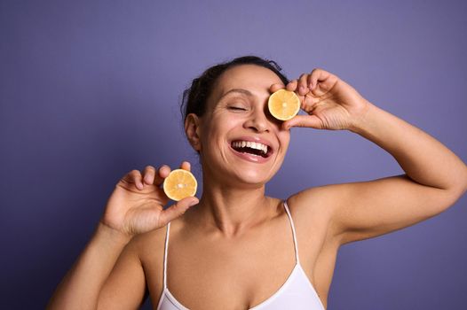 Beauty portrait of attractive cheerful smiling young woman with curly dark hair wearing white top isolated on bright purple background holding lemon halves and covering her eyes with it. copy ad space