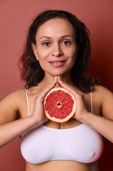 Beauty portrait of an attractive smiling middle aged beautiful woman with dark curly hair hair, wearing white underwear, isolated on bright coral background, holding half of fresh juicy red grapefruit