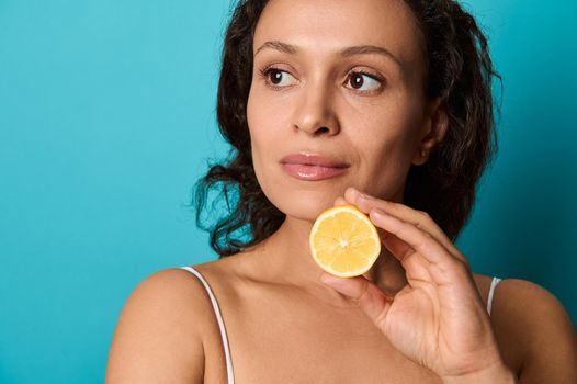 Close-up confident portrait of a natural beauty charming attractive elegant woman with curly dark hair isolated on bright blue background holding lemon half. Health care, skin treatment, body care.