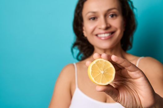 Beauty portrait of attractive cheerful smiling young woman with curly dark hair in white top isolated on bright blue background holding halves of lemon and covering her eyes with it. Copy ad space
