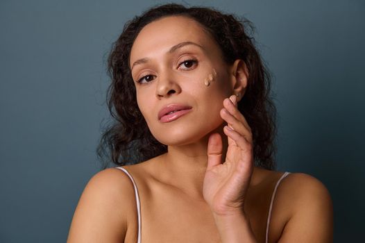 Portrait of beautiful elegant woman with drops of fluid corrective foundation on her cheek, looking confidently at camera, isolated on grey-blue background with copy space. Makeup, skin care concept