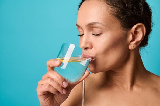 Headshot of a beautiful woman with fresh healthy skin drinking lemon water from transparent glass, isolated over bright blue background with copy ad space. Healthy eating and lifestyle concept