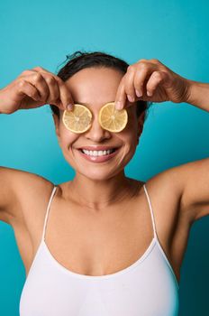 Natural beauty young attractive woman with healthy perfect fresh skin covers her eyes with half a lemon, smiles beautiful white toothy smile isolated on bright blue background with copy space for ads
