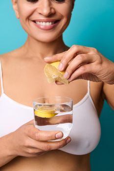 Soft focus in the hands of blurred pretty beautiful woman smiling toothy smile squeezing a fresh lemon juice into a glass with water, preparing lemon water, isolated on blue background with copy space