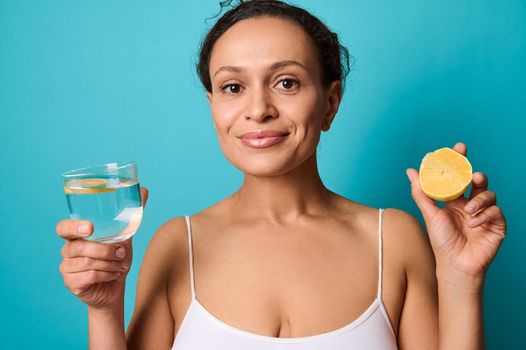 Beautiful middle aged woman holding a half of fresh juicy yellow lemon and a glass with water in her hands, smiling cutely looking at camera isolated over blue background with copy space for ads