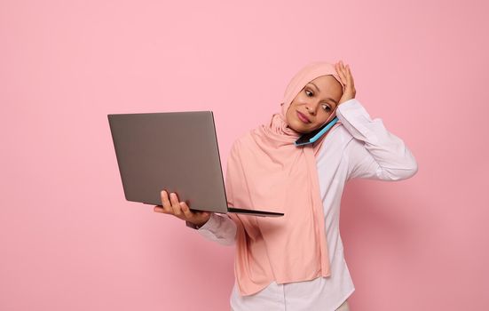 Portrait of puzzled Muslim Arab woman in pink hijab, with laptop, talking on the phone, holding her head because of multitasking, looking down with sad expression, on colored background copy space