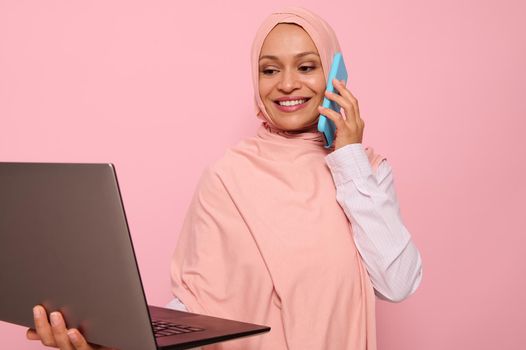 Close-up portrait of a gorgeous Muslim Arab woman in pink hijab working on laptop, isolated on colored background with copy space. Successful programmer, IT female worker, start-up, business lady