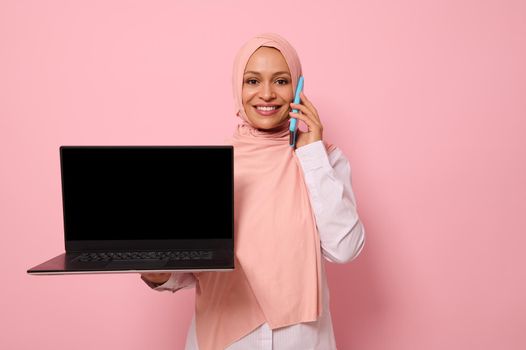 Attractive successful Arab Muslim woman in pink hijab talks on mobile phone, smiles with toothy smile looking at the camera and shows the empty blank black monitor screen with copy space to the camera