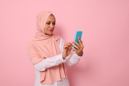 Isolated portrait of confident Arab Muslim mature pretty woman in strict religious outfit and covered head in pink hijab texting message on a mobile phone in her hands, colored background, copy space