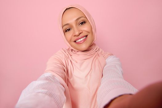 Close-up portrait of Arab Muslim woman wearing traditional religious islamic outfit, pink hijab, holding smartphone in outstretched arms and smiles with toothy smile making a selfie, self-portrait