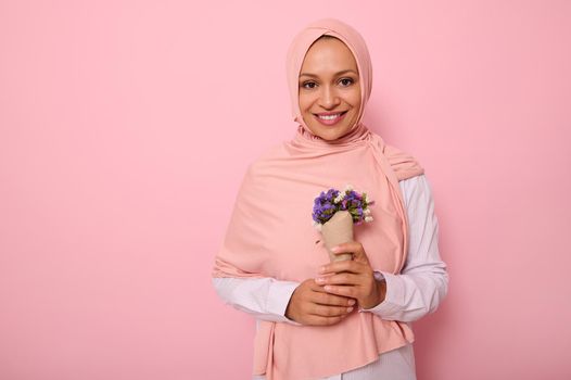 Isolated portrait on colored background of a beautiful Muslim Arab woman in pink hijab, holding a bouquet of wildflowers in purple shades, wrapped in craft paper, smiles at camera with a toothy smile