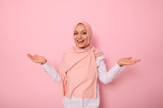 Pleasantly surprised gorgeous Arabic muslim woman with covered head in pink hijab joyfully looking at camera smiling beautiful toothy smile while standing against pink background, hands turned palm up