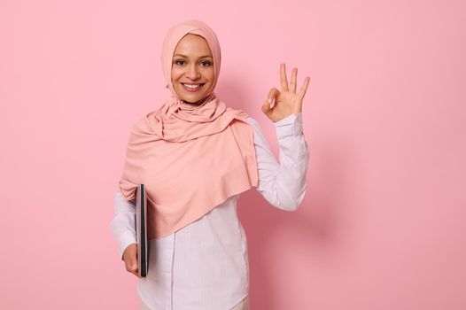 Gorgeous young Muslim woman of Middle Eastern ethnicity in hijab holds a laptop under arms and showing OK sign with fingers, smiles with toothy smile looking at camera, pink background copy space.