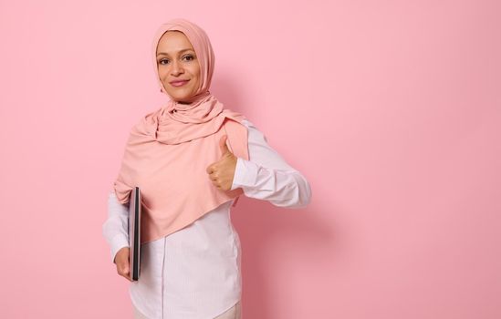 Confident portrait of a successful gorgeous Arab Muslim woman in pink hijab and strict casual attire showing a thumb up, looking at camera, posing against colored pastel background with copy space