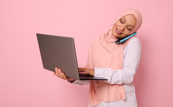 Concept of a busy Arab muslim business woman, freelancer in hijab, talking on a mobile phone while working on a laptop at the same time. Isolated on pink background with copy space. Confident portrait