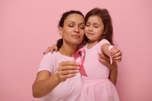 Young mixed race woman mother hugs little girl in pink outfits and displays pink satin ribbon, symbol of Breast Cancer Day in October Month. Support for cancer patients, survivors. Colored background