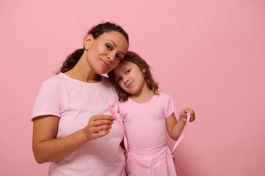 Cute baby girl , leans on her mother shoulder,both dressed in pink color outfit, hold pink satin ribbons, cute smile looking at camera . World Breast Cancer Awareness Day. Fight Cancer. Copy space