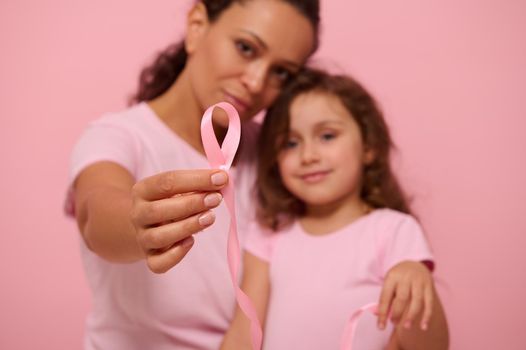 Focus on a long pink satin ribbon with one endless end in the hands of blurred two generations of pretty women, mother and daughter, expressing support and solidarity for cancer patients and survivors