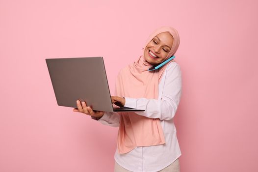 Successful Muslim Arab gorgeous woman in hijab, dressed business smart casual talks on mobile phone , smiles looking at the monitor screen while typing text on a laptop. Copy space, pink background