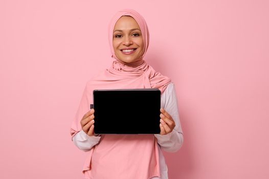 Arab Muslim beautiful woman wearing pink hijab, smiling toothy smile looking at camera, standing against pink background with copy space and showing the empty blank black screen of a digital tablet