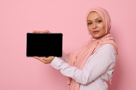 Confident Muslim Arab beautiful woman with covered head in hijab holds horizontally a digital tablet and shows its blank screen looking at the camera, isolated on pink background with copy space