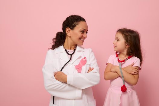 Beautiful woman doctor and cute baby girl looking at each other, wearing pink ribbon, symbol of Breast Cancer Awareness Day, standing with crossed arms on chest on colored background with copy space