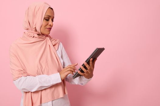 Beautiful Arab Muslim woman with covered head in pink hijab posing against a colored background with a digital tablet in hands, copy space for text. Advertising isolated portrait
