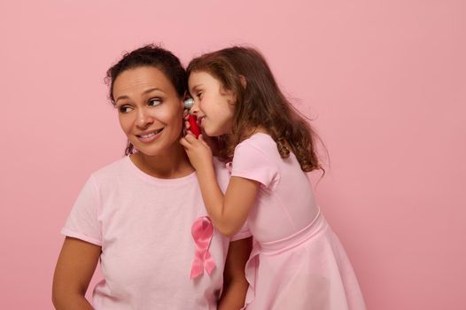 Little girl playing doctor with her mother. Beautiful mixed race woman with baby girl in pink with pink ribbon, symbol of International Breast Cancer Awareness Day. Medical Educational concept
