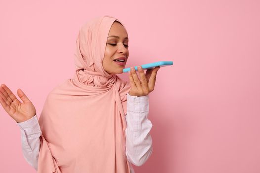 Muslim Arab gorgeous beautiful woman with covered head in pink hijab recording a voice message on her smartphone. Isolated portrait on pink pastel background, copy space for advertising