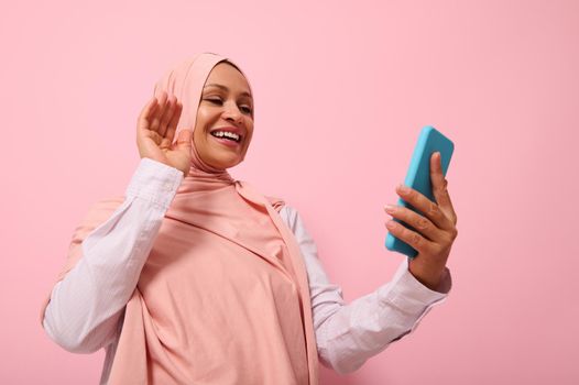 Muslim woman of Middle Eastern or Arab ethnicity with covered head in pink hijab having a video conference on mobile phone, greeting her interlocutor during video call, pink background copy space