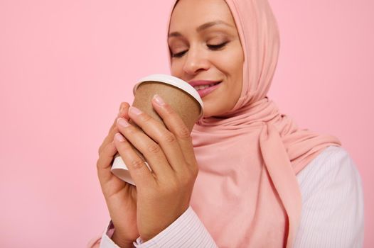 Arabic Muslim pretty woman with covered head in hijab drinking hot drink, tea or coffee from disposable cardboard takeaway cup, standing three quarters against colored background, copy space. Close-up