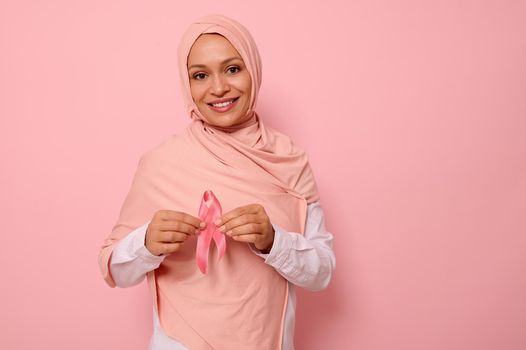 Friendly and solidar Arab Muslim woman in a pink hijab holds a pink ribbon to her chest levels for a breast cancer campaign, promoting cancer awareness. Pink October month , women's health concept