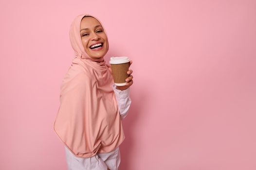 Charming Muslim woman with covered head in pink hijab holds disposable cardboard takeaway cup, smiles toothy smile, looking at camera, standing three quarters against colored background, copy space