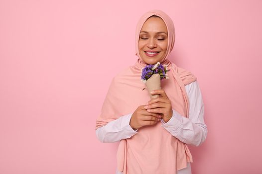 Portrait on pink background of beautiful Muslim Arab woman in hijab with closed eyes and charming smile enjoying the smelling of meadow flowers wrapped in craft paper. International Women's Day concept