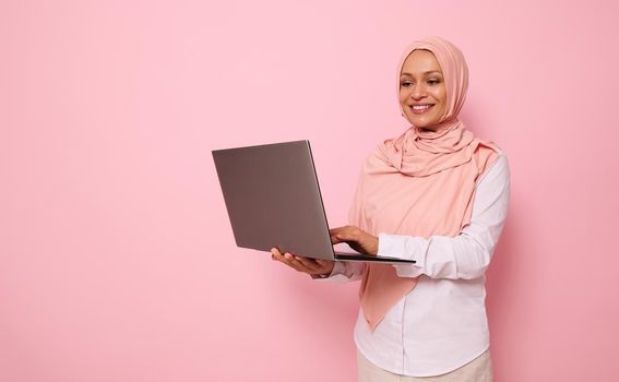 Isolated portrait on pink background with space for text of a successful middle aged Muslim beautiful woman of Middle Eastern ethnicity wearing hijab and dressed smart casual outfit working on laptop
