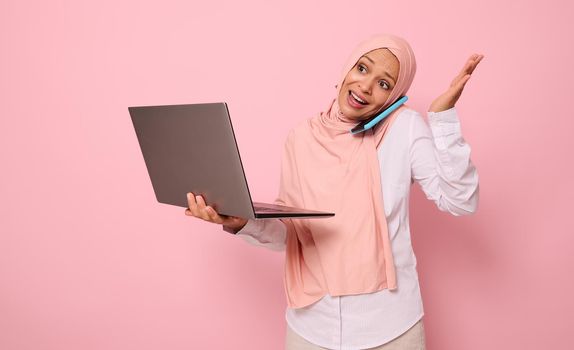 Busy and puzzled Muslim Arab gorgeous pretty woman in hijab, dressed business smart casual holds a laptop and talks on mobile phone , gestures with hands. Isolated over pink background with copy space