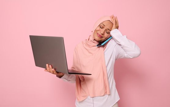 Puzzled Muslim woman in hijab, with laptop, talking on the phone, holding her head because of multitasking. Concept of a busy young Arab business lady, freelancer, office worker on colored background